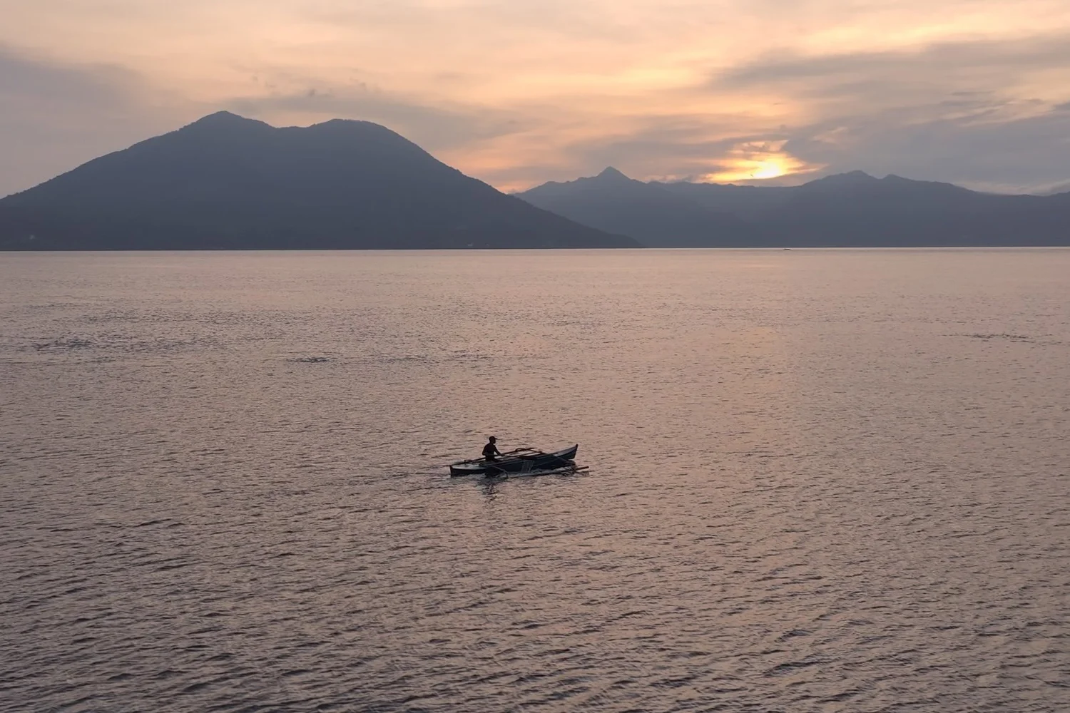 Fisherman in an outrigger canoe at sunset with mountains in the distance